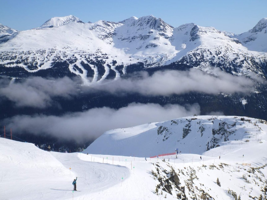 Fog hangs over a ski field dotted with skiers, with snow-capped mountains in the background.