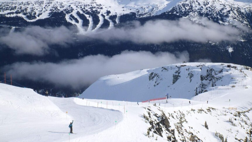 Fog hangs over a ski field dotted with skiers, with snow-capped mountains in the background.