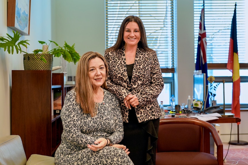 A seated Tammy Tyrrell and a standing Jacqui Lambie post in a Senate office. Both have long hair that's not tied back. 