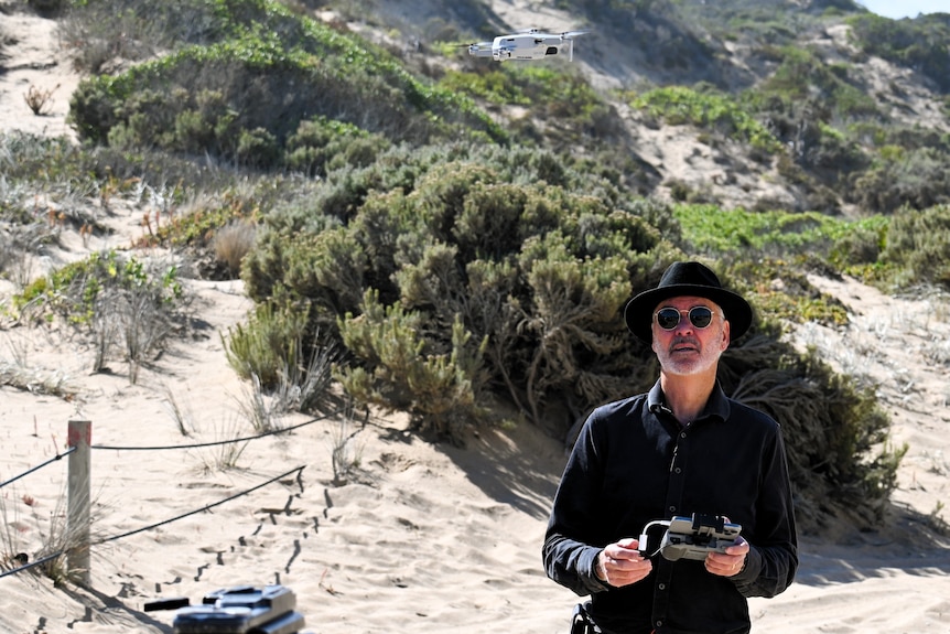 A man holding a remote control stares upwards at a drone, sandhills in the background