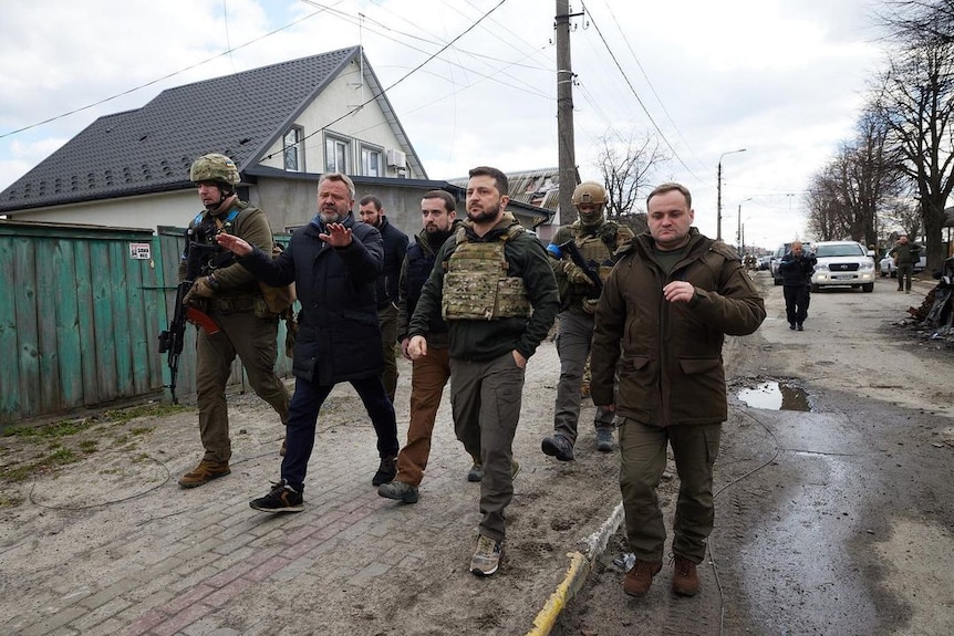 President Zelenskyy in a bullet proof vest walks through a town, surrounded by soldiers and other men 