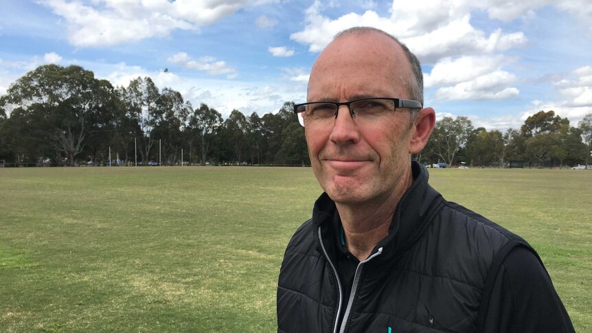 A man with a balding head standing on a green sports oval.