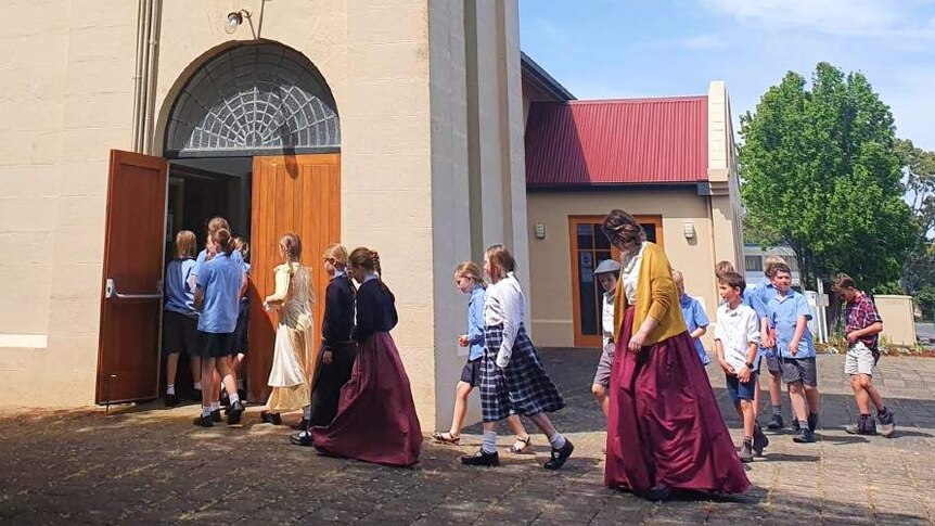 Young school children walk into a large beige church building surrounded by leafy green trees.
