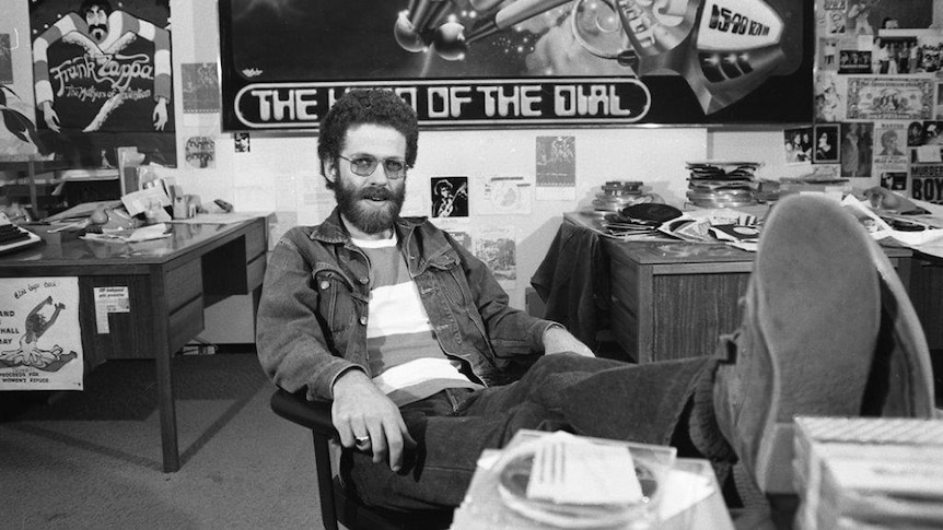 Black and white photo of Double Jay announcer Chris Winter with his feet on his desk in an office full of records and posters