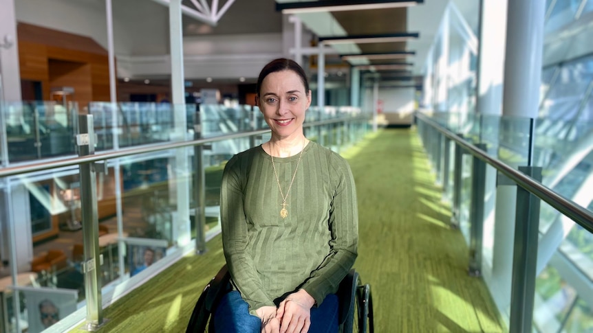 woman smiles brightly at the camera, she wears a green shirt and is seated in a wheelchair