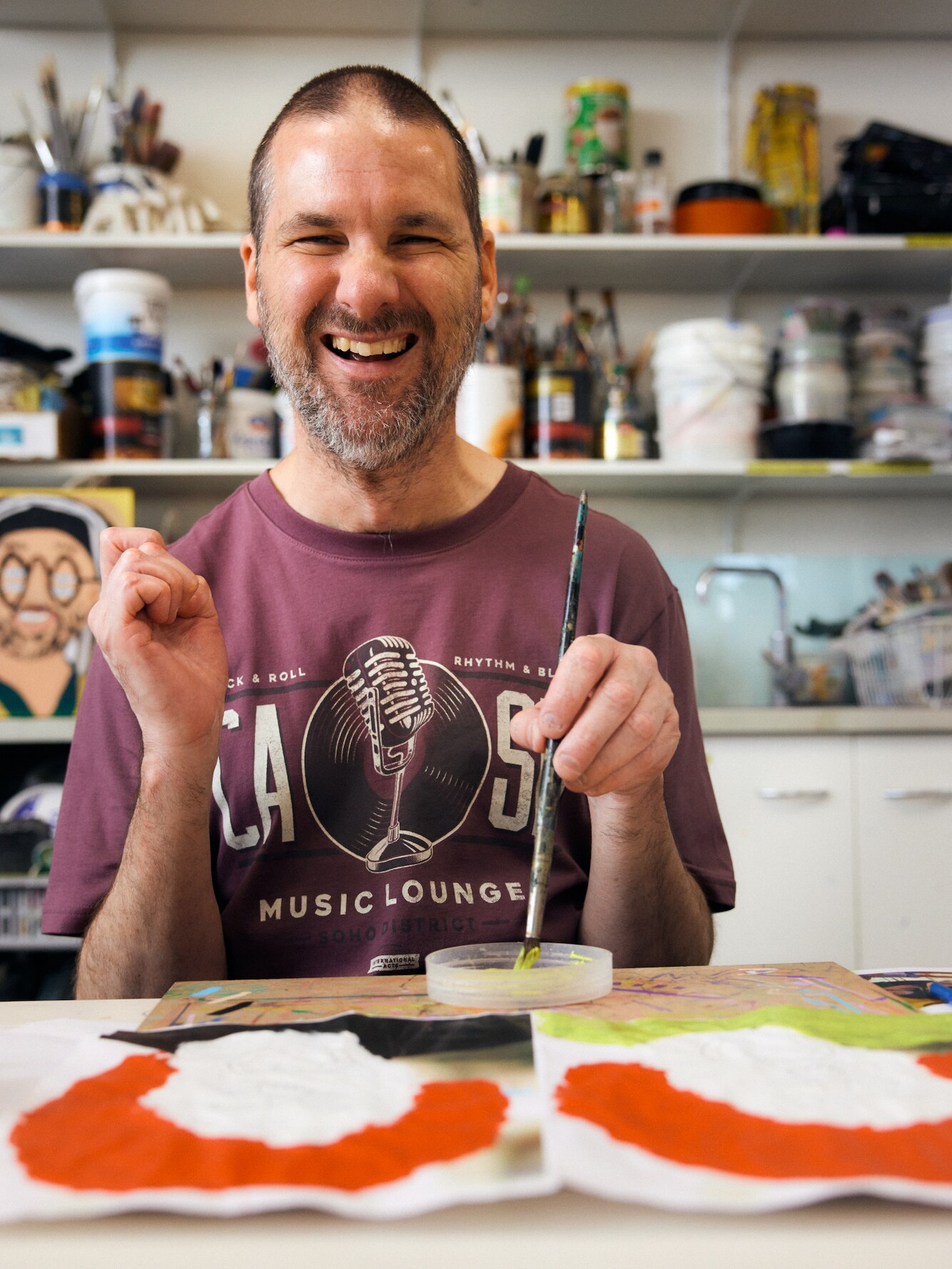 Photo of the artist smiling at the camera, sitting in his art studio. He wears a maroon t-shirt and holds a paintrbrush 