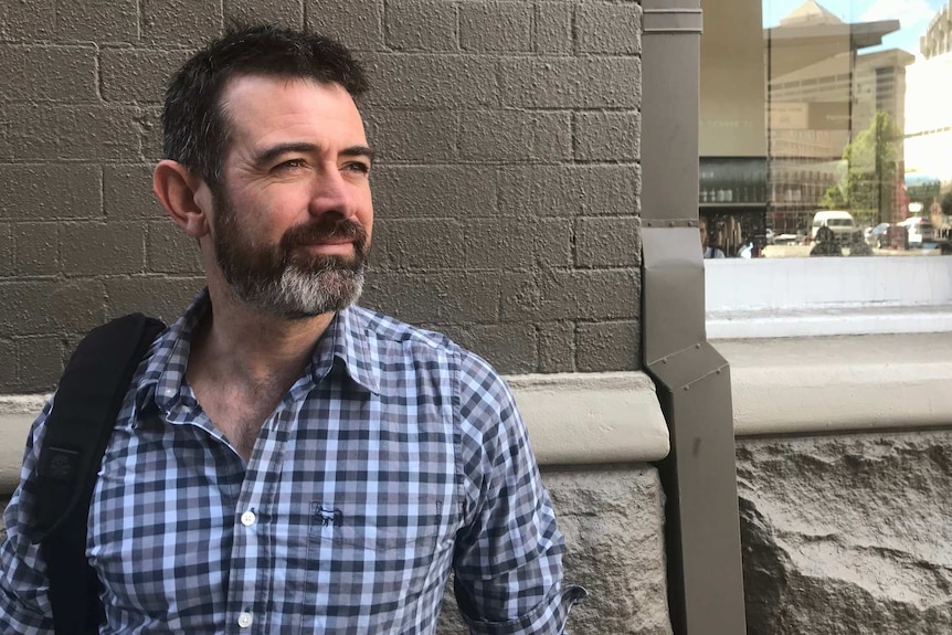 Gearoid Towey, wearing a checked blue shirt, stands in front of a grey brick wall.