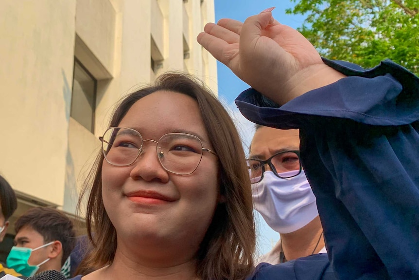 A young Thai woman in a blue top and wire-rimmed glasses does a three-fingered salute
