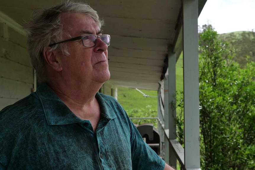 An older man wearing glasses stands on a wooden deck looking out.