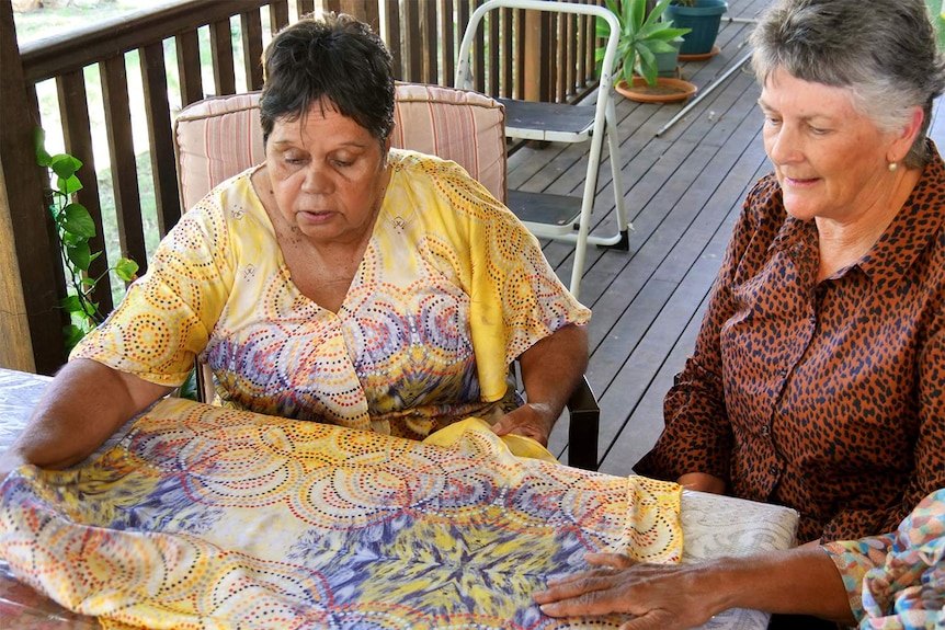 Two ladies looking at a yellow silk scarf with a colourful Aboriginal dot painting design on it.