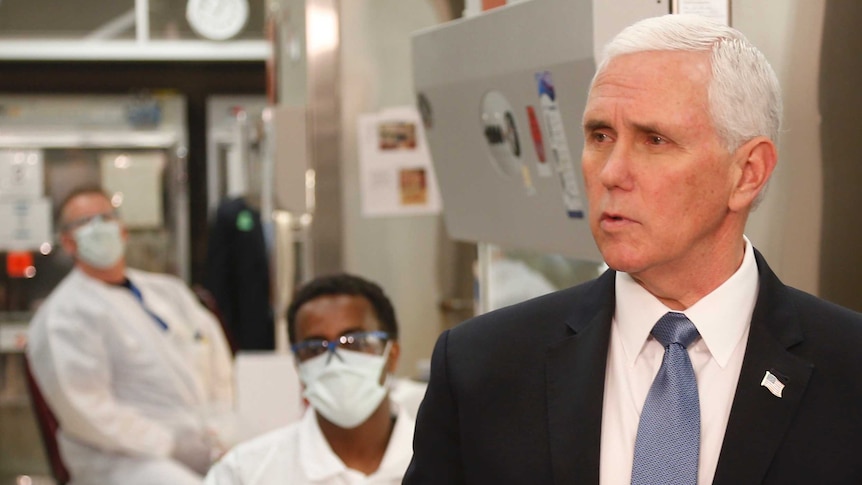 Mike Pence speaks while two people sit behind him in lab coats and face masks.