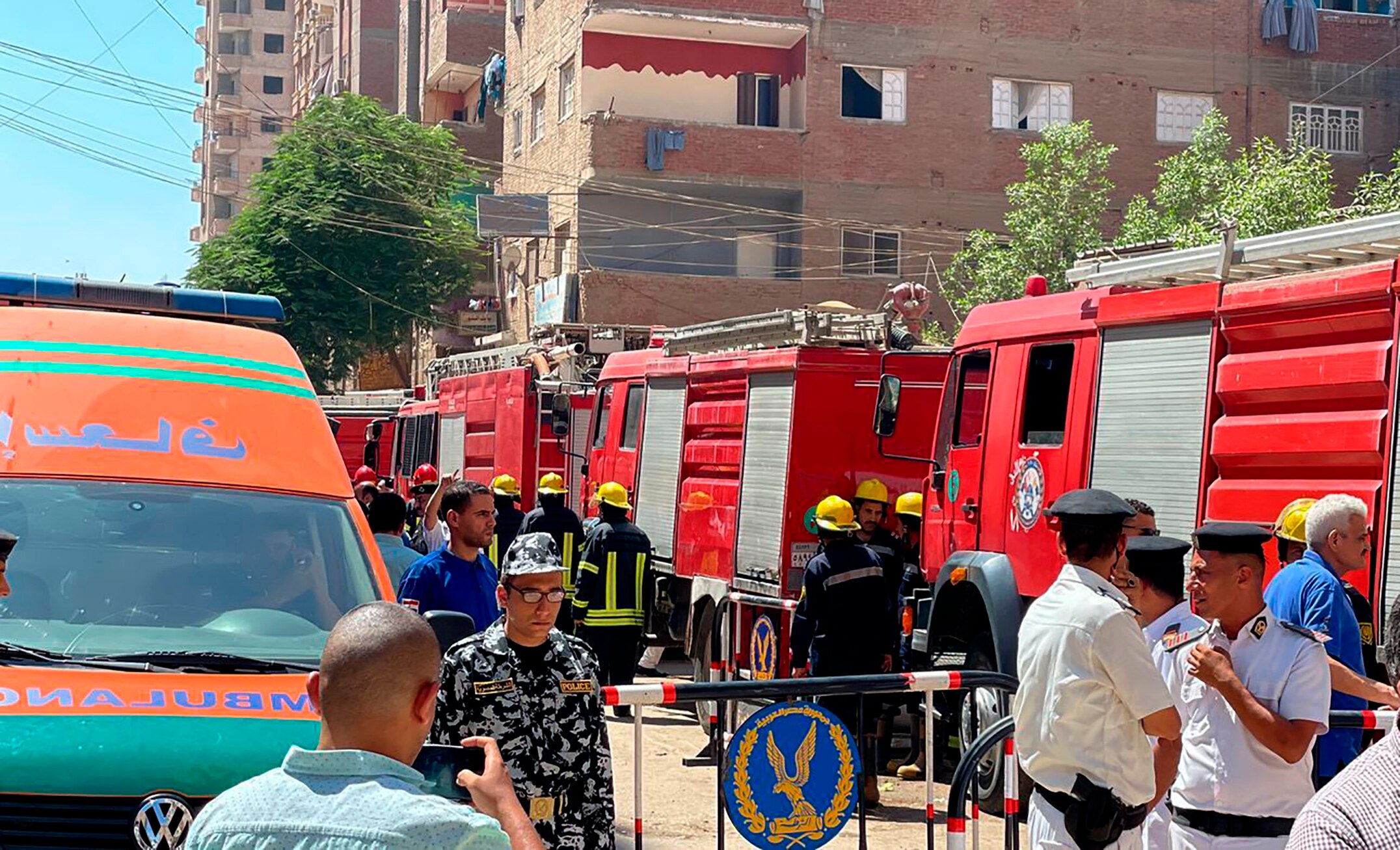 Emergency personnel work at the site of a church fire, with four fire trucks parked in a narrow street with many onlookers.