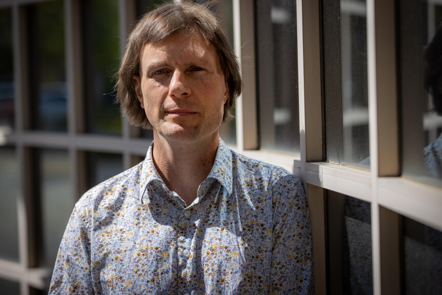 A man with straight brown hair wearing a floral shirt stands next to windows
