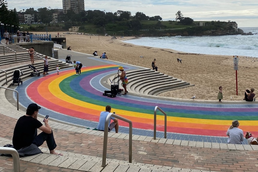 People sitting around an ampitheatre near the ocean, with a rainbow painted on the walkway.
