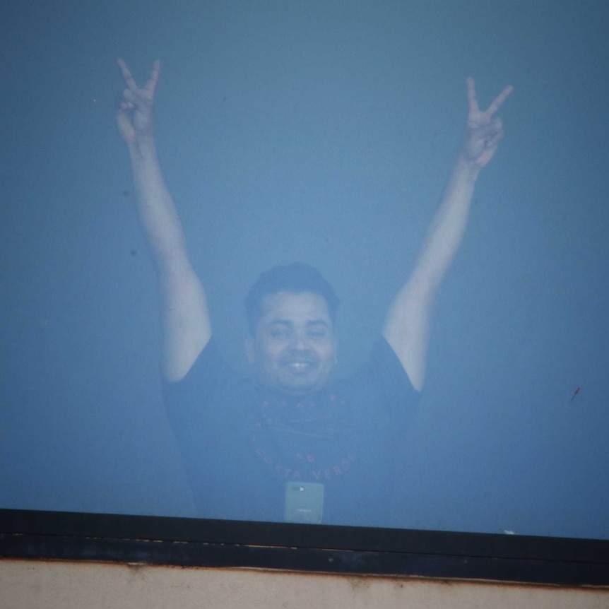 Man with his arms raised making a V sign behind a hotel window.