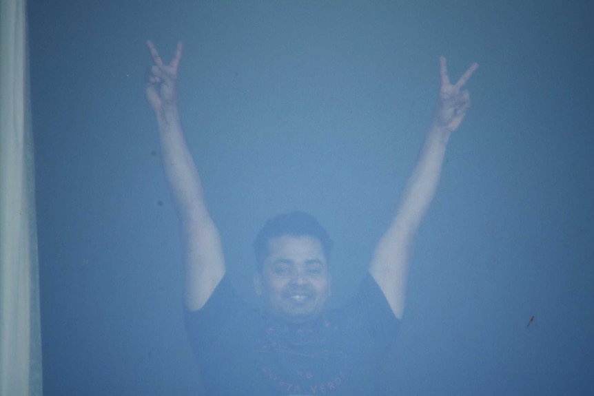 Man with his arms raised making a V sign behind a hotel window.