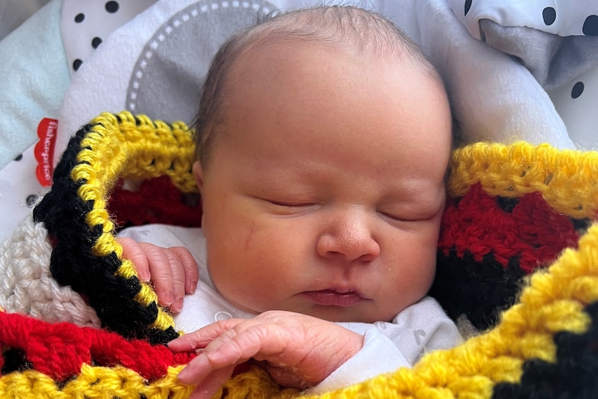 A close up of a baby's face. He is wrapped in a crocheted blanket in black, yellow, red and white.