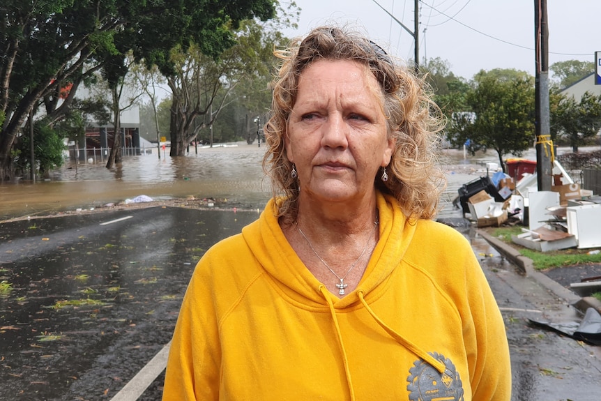 woman with blonde curly hair, wearing a yellow jumper, standing on a road with flood waters and piles of rubbish behind her