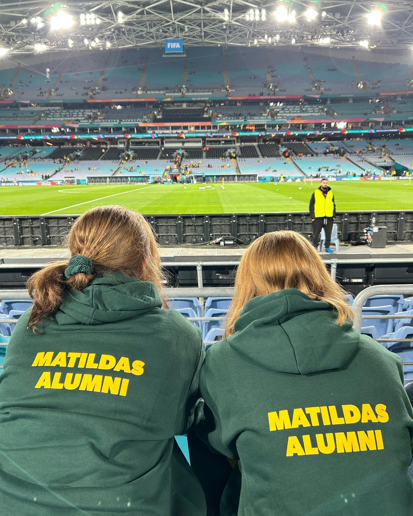 Two women wearing green and gold Matildas Alumni jumpers at a football stadium