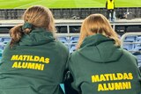 Two women wearing green and gold Matildas Alumni jumpers at a football stadium