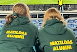 Two women wearing green and gold Matildas Alumni jumpers at a football stadium