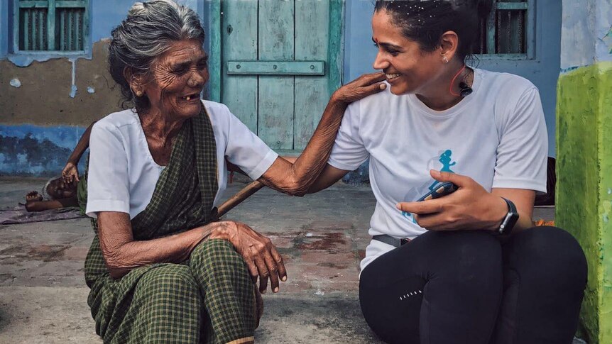 Srishti Bakshi (right) sits with an older Indian woman (left) in front of a blue house. They are facing each other and smiling