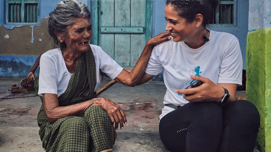 Srishti Bakshi (right) sits with an older Indian woman (left) in front of a blue house. They are facing each other and smiling