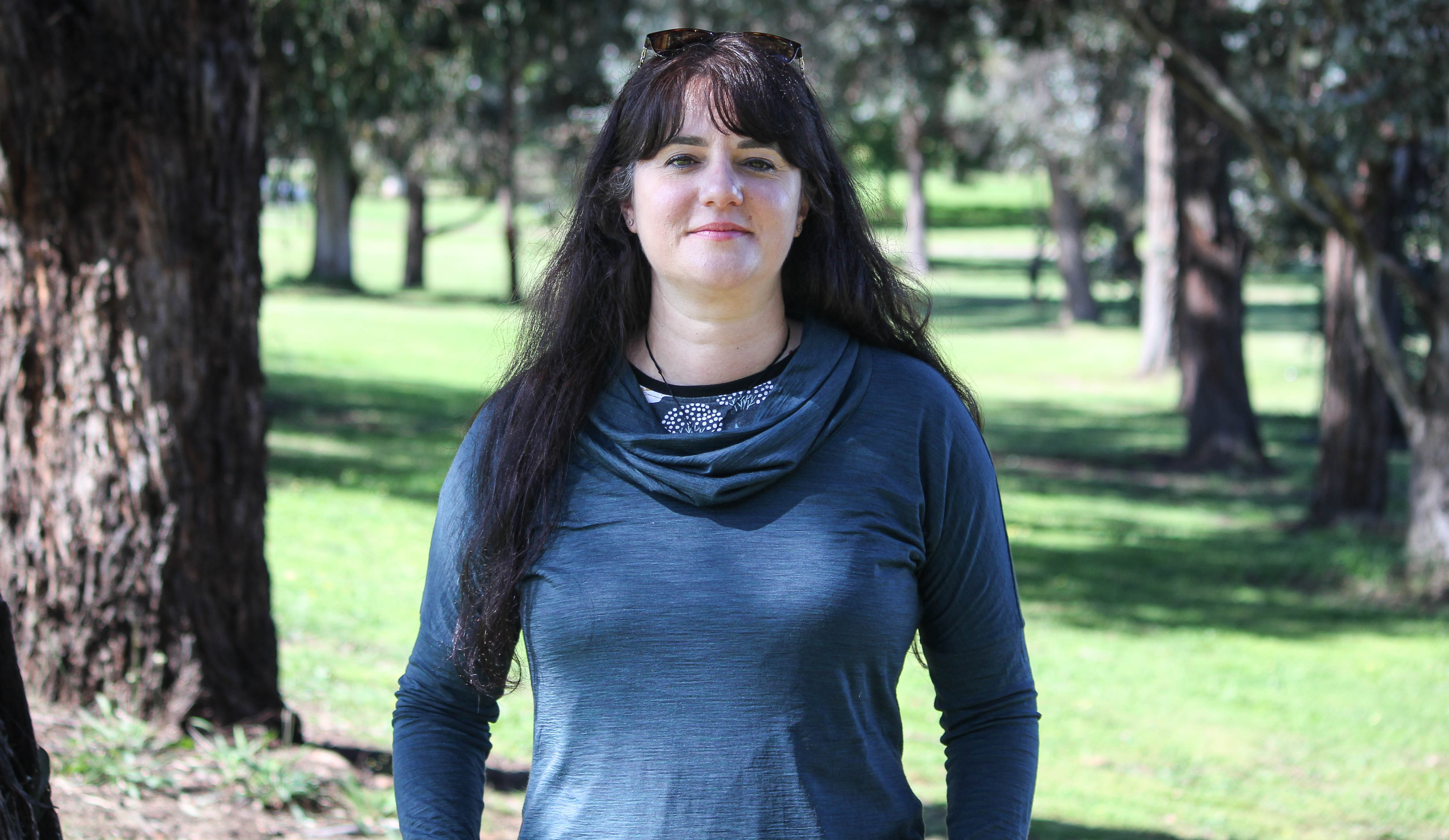 Woman with long brown hair and blue shirt stands in a park with trees in the background.