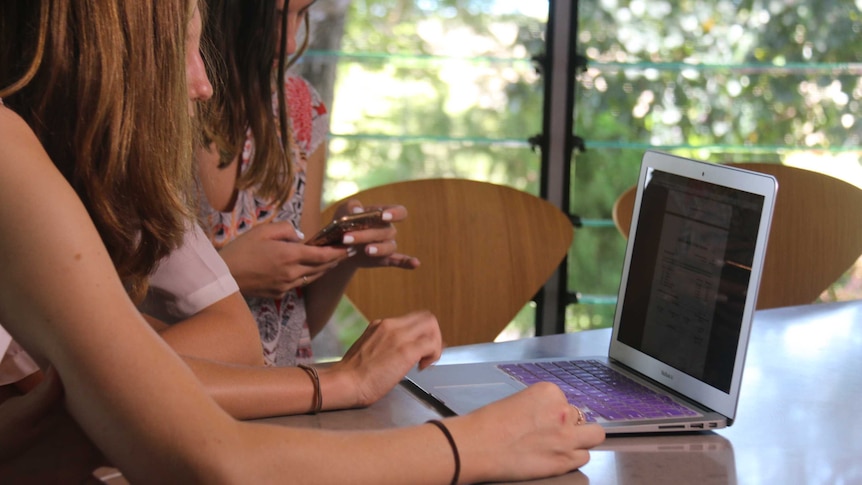 Unidentifiable teenage girls look at a laptop on a bench top while another looks at her iPhone.