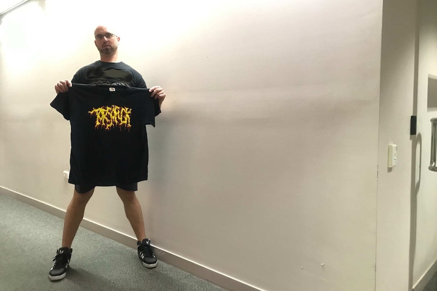 Wide angle photo of a man wearing a black metal t-shirt, holding another t-shirt standing in front of a white wall