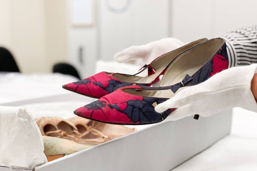 A pair of black and red high-heeled shoes held in gloved hands.