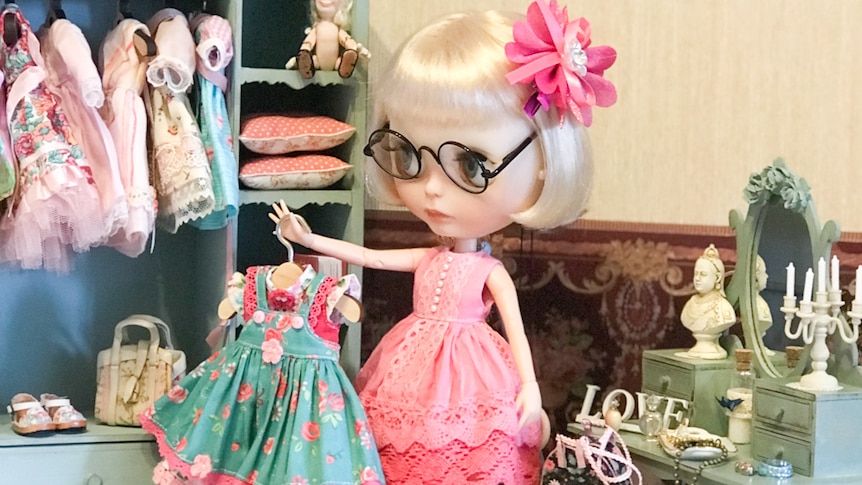Doll holding a dress stands in front of miniature wardrobe