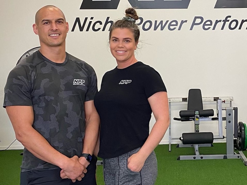 A man and a woman in exercise gear stand in a gym and smile at the camera