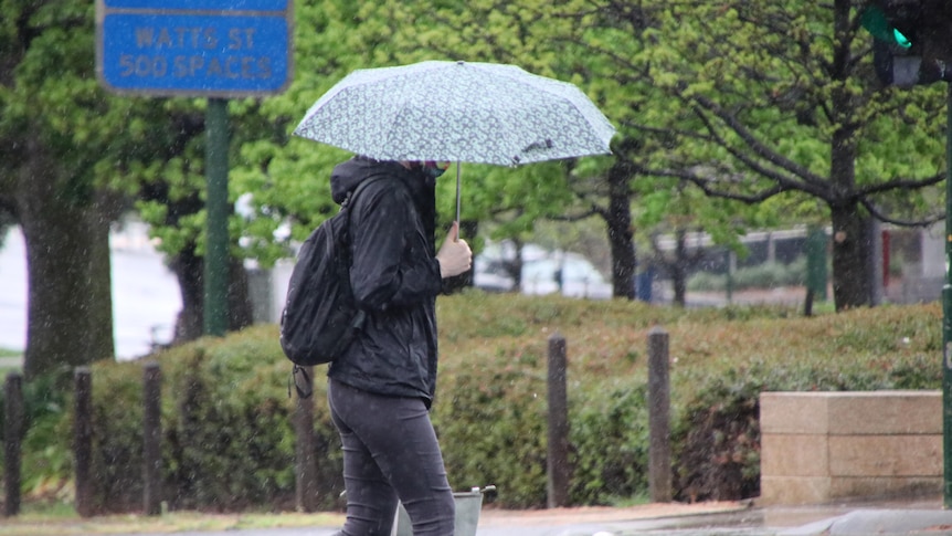 A person wearing a face mask walks holding an umbrella on a rainy day.