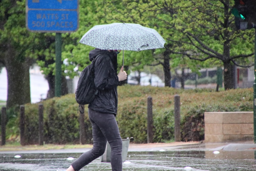 A person wearing a face mask walks holding an umbrella on a rainy day.