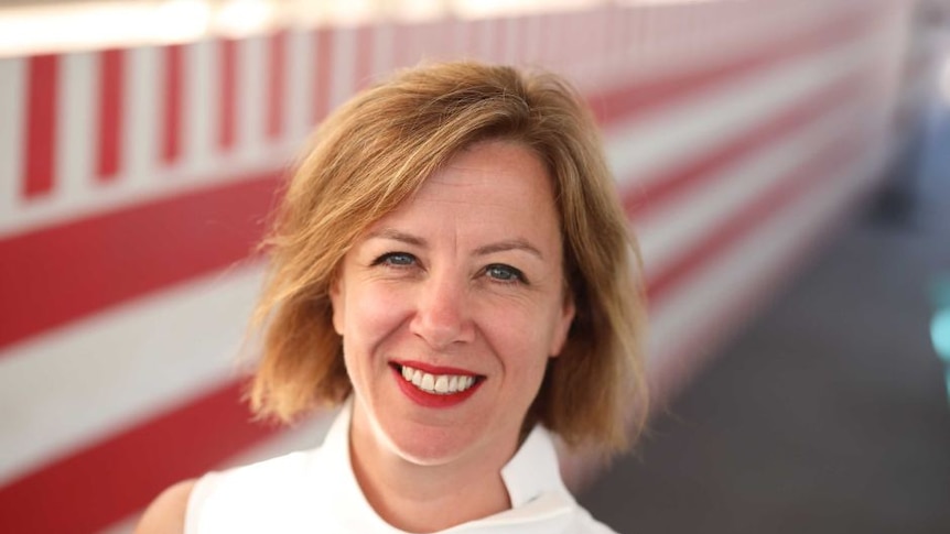 A blonde-haired white woman wearing a white sleeveless top smiling at the camera against a red and white background