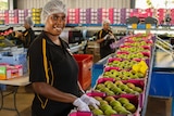 A smiling woman wearing a hair net and work uniform packs green mangoes into boxes with gloved hands.