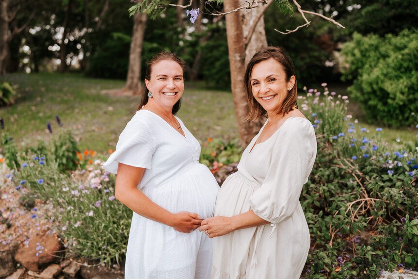 Sophie and Jasmina wearing white dresses with large pregnant bellies, smiling, garden behind.