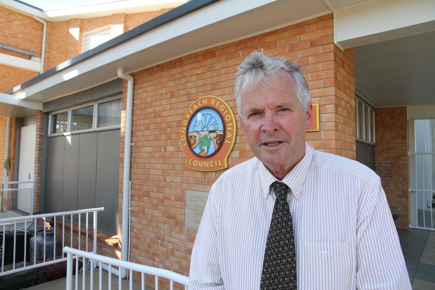 A man in a shirt and tie stands in front of a brick building with a colourful regional council logo on it.