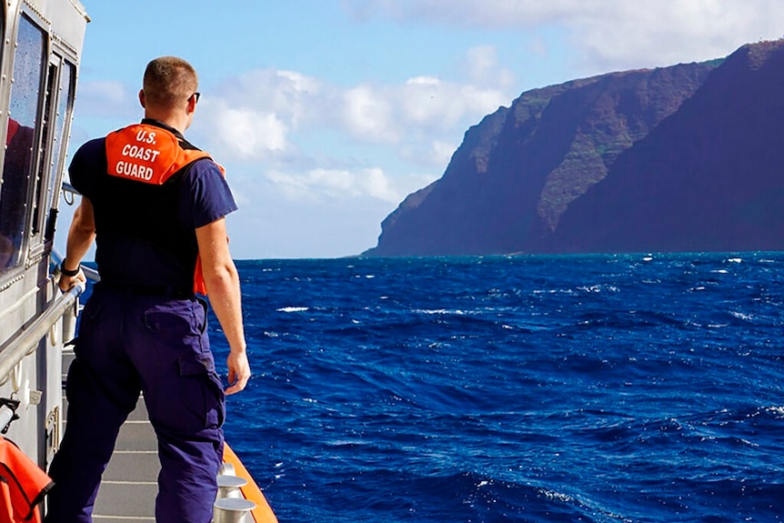 Looking from behind, you few a US Coast Guard holding onto the side of a coast guard vessel in rich blue waters.