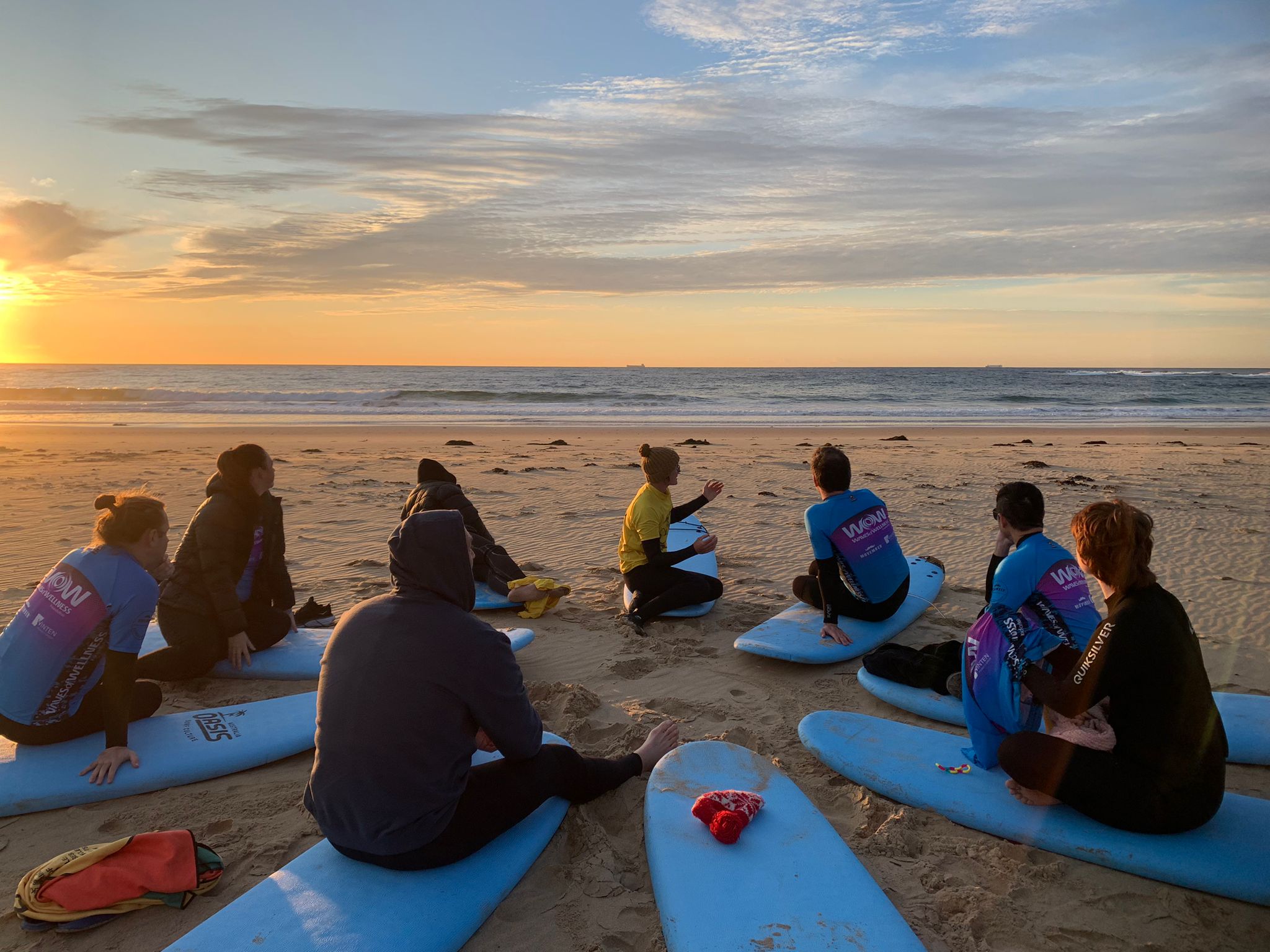 A group of people sit in a circle on surfboards, on a beach at sunrise.