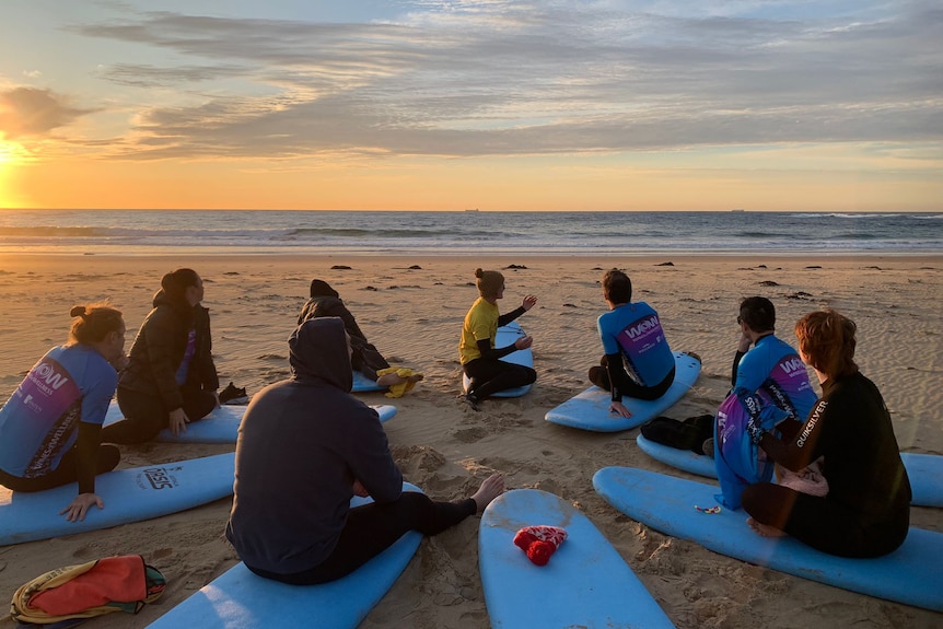A group of people sit in a circle on surfboards, on a beach at sunrise.