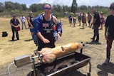 A man draped in an Australian flag stands over a spit-roasting pig.