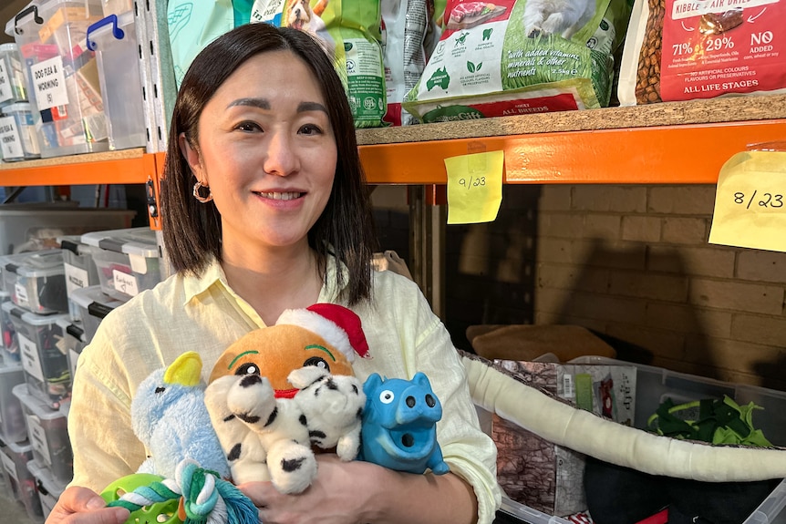 A woman with dark shoulder length hair and yellow shirt, holds a bunch of pet toys in a warehouse. 