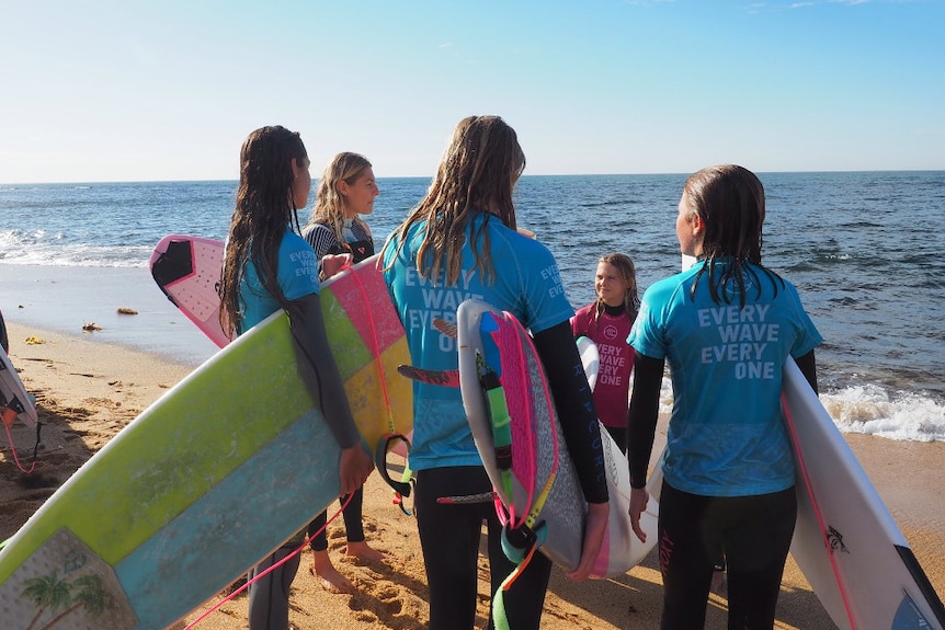 Four female surfers stand in a circle on the beach with their boards.