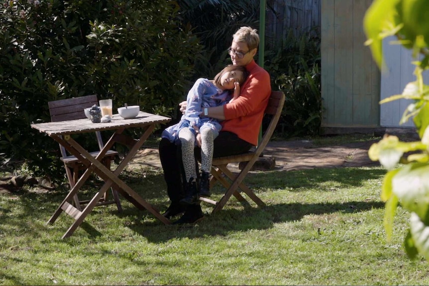 Kristine Rowe sits at an outdoor table, her daughter sits on her knee and leans against her while tiredly wiping her eye.