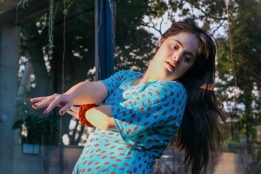 Brown-haired woman with Down syndrome dances in afternoon light on the back lawn of her home, wearing a sky blue and red dress