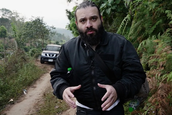 A man with a beard and a black jacket standing in a tropical forest area