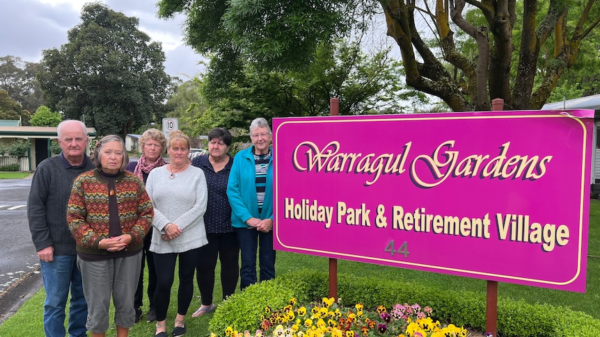 Six residents stand by a pink Warragul Gardens sign above a pretty flower bed.