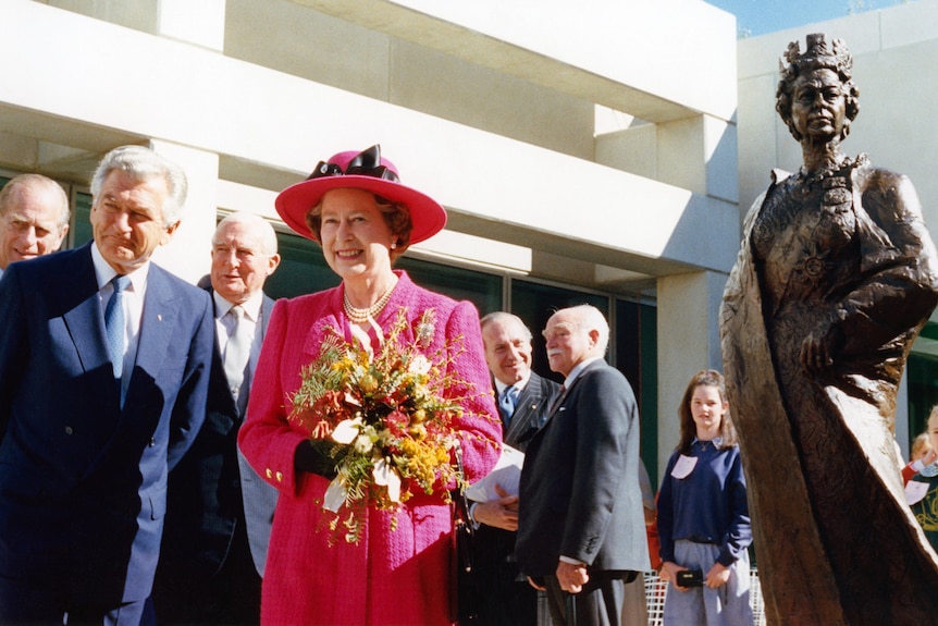 Queen Elizabeth II holds a bouquet of flowers while standing next to a statue of herself.